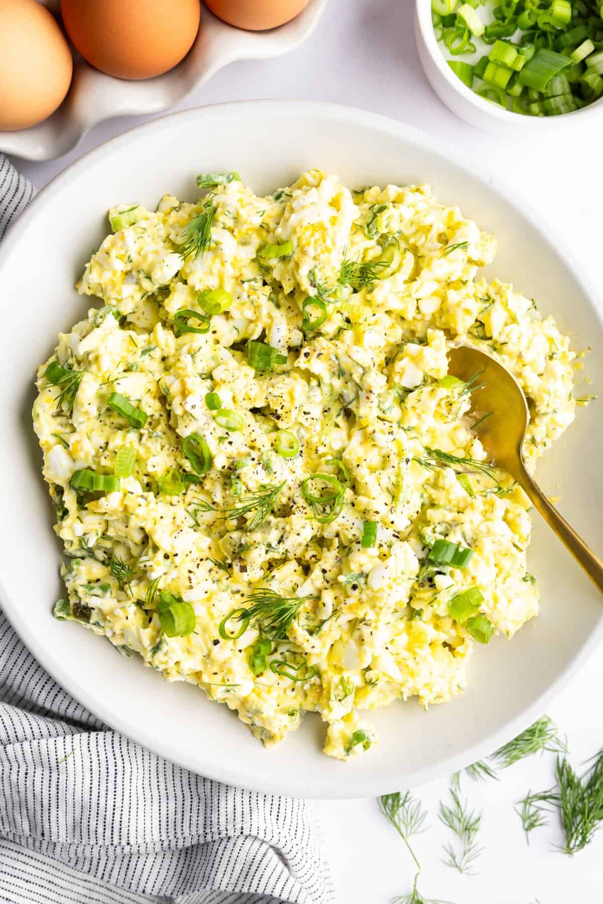 A bowl of egg salad garnished with chopped green onions and dill, placed next to whole eggs, sliced green onions, and a striped cloth.