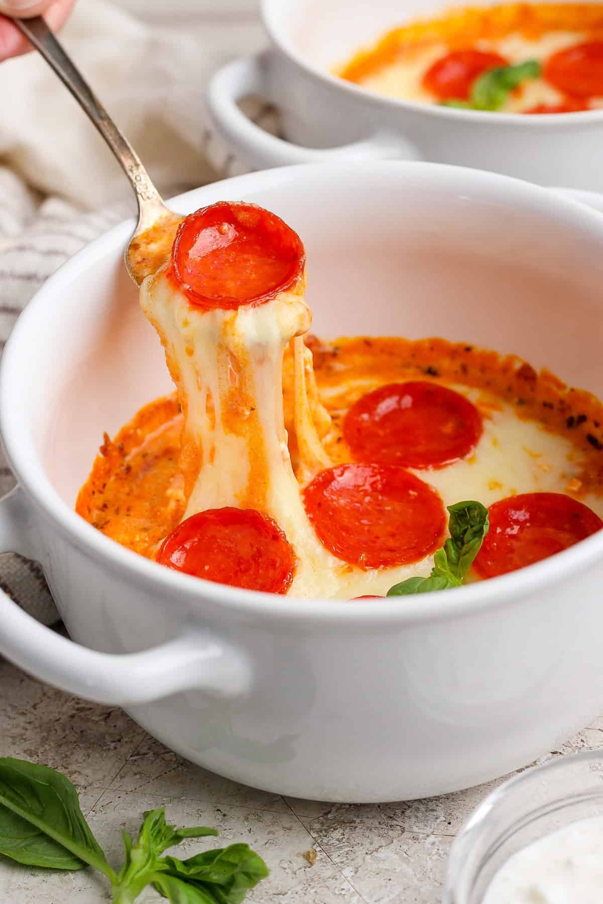 Egy kanál felemeli a sajtos pepperoni pizza mártást egy fehér tálból. A friss bazsalikom levelek láthatók a közelben.