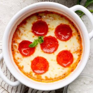Sült étel olvasztott sajttal és hat pepperoni szelet, bazsalikommal díszítve, egy fehér kerámia tálban egy texturált felületen.