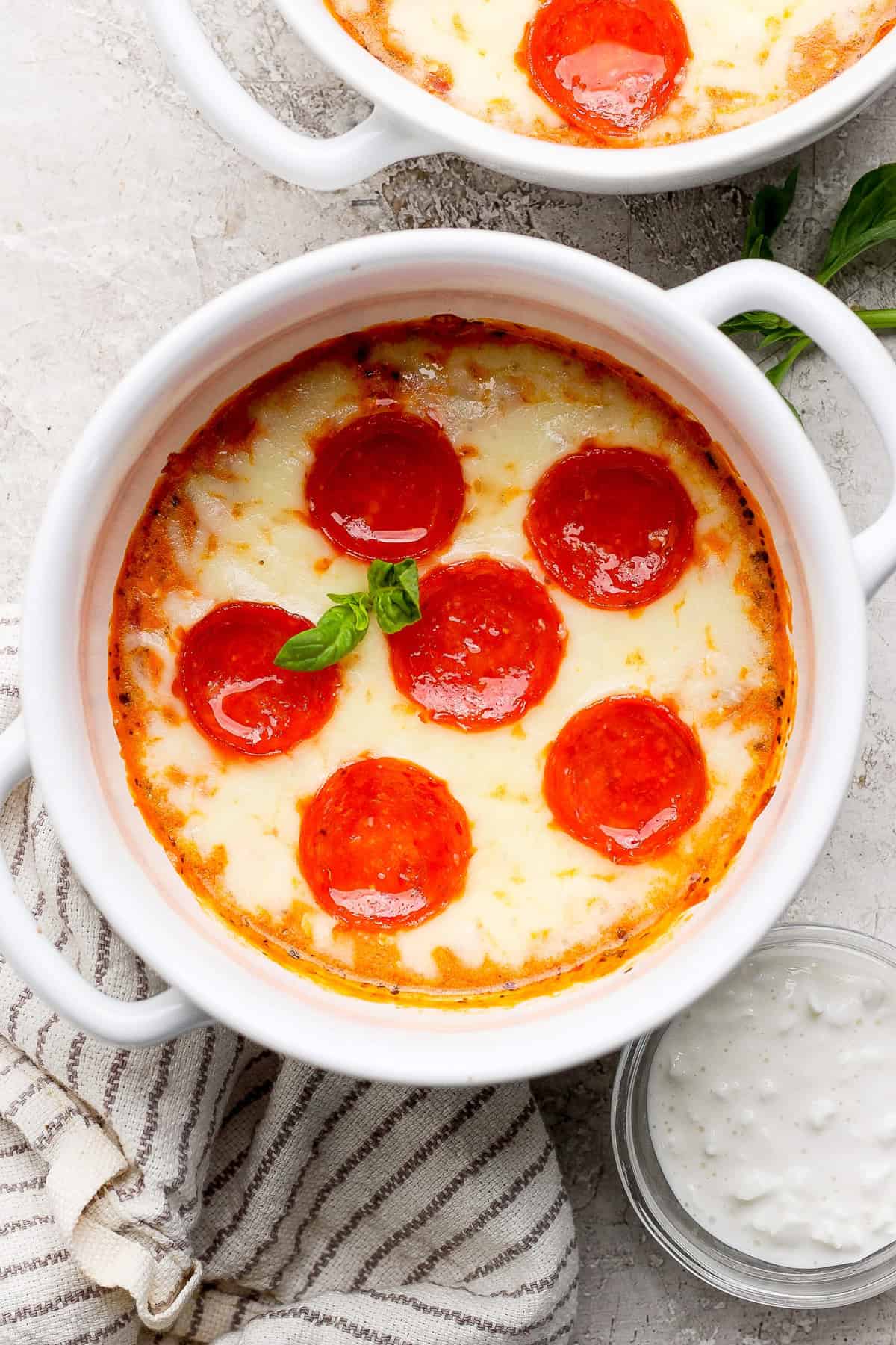 Egy fehér tál sült pepperoni és sajtmártással, bazsalikommal díszítve, egy kis tál mártó mártás melletti ruhára helyezve.