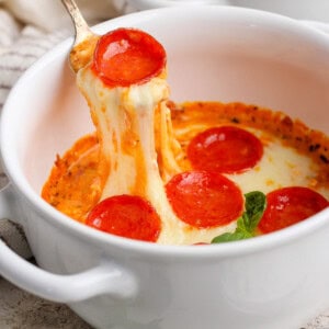 Egy kanál felemeli a sajtos pepperoni pizza mártást egy fehér tálból. A friss bazsalikom levelek láthatók a közelben.