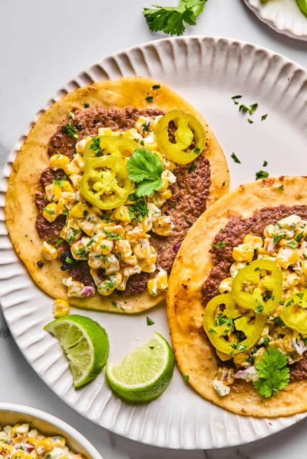 Két tacos egy fehér tányéron, amelyet őrölt hús, kukoricasaláta, pácolt jalapeños, koriander, és mész ékkel tálalva.