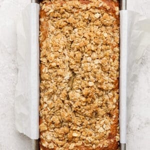 Loaf of banana bread with oat crumble topping in a parchment-lined metal baking pan on a light textured surface.