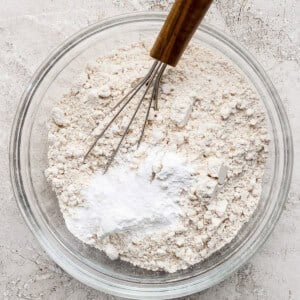 A glass bowl contains a mixture of flour and baking powder, with a metal whisk resting inside, placed on a textured gray surface.