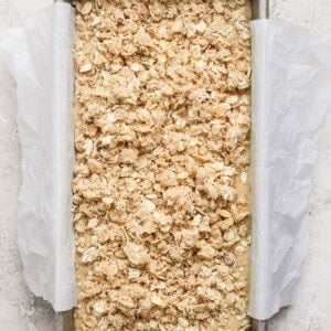 A rectangular baking dish lined with parchment paper contains an unbaked oat crumble mixture on a light textured surface.