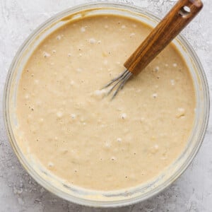 A glass bowl filled with thick, creamy batter is placed on a textured gray surface. A wooden-handled whisk rests inside the bowl.