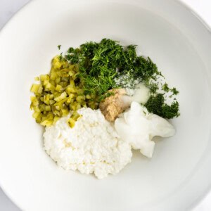 White bowl containing cottage cheese, chopped pickles, fresh dill, minced garlic, salt, and sour cream.