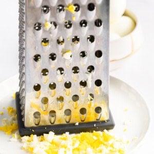 A metal cheese grater on a white plate with finely grated egg, alongside a bowl containing whole boiled eggs.
