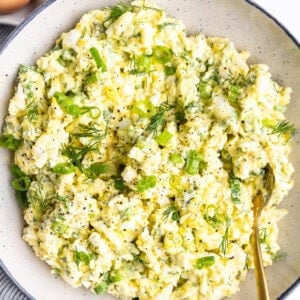 A bowl of egg salad with chopped green onions and dill, seasoned with black pepper. Three whole eggs are in the background.