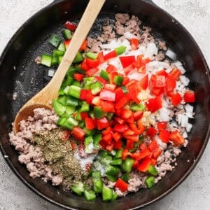 A cast iron skillet with ground meat, chopped red and green bell peppers, onions, garlic, and herbs, with a wooden spoon.