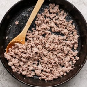 Ground meat cooking in a black cast-iron skillet with a wooden spoon on a textured white surface.