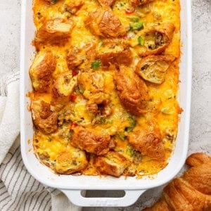 A casserole dish filled with a baked croissant breakfast casserole topped with melted cheese and diced vegetables on a textured surface. A white striped cloth is partially visible.