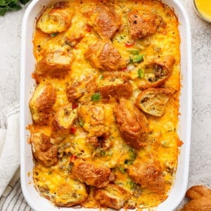 A baked casserole in a white dish, topped with melted cheese and croissant pieces. Two glasses of orange juice and fresh herbs are nearby.