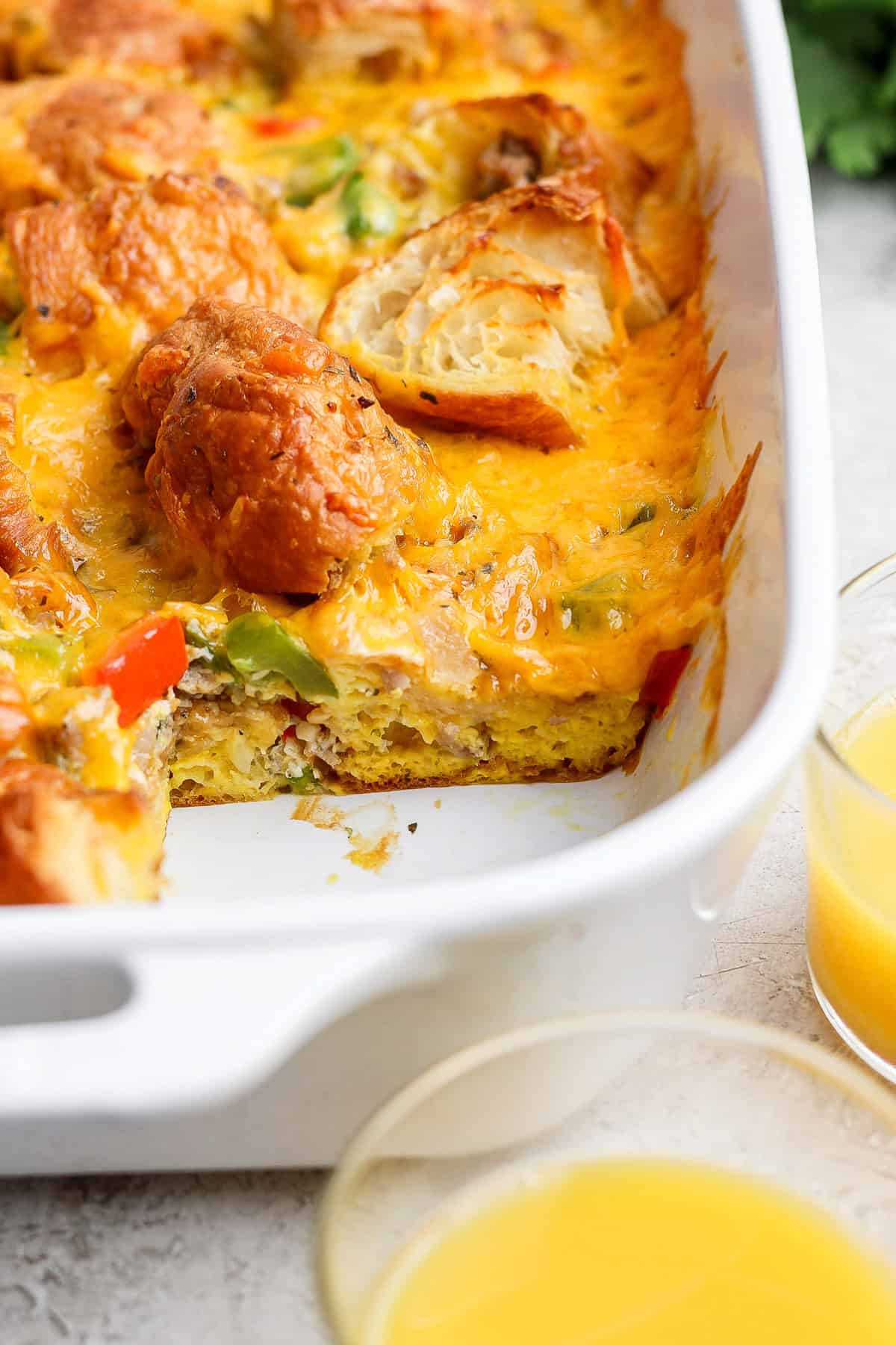 A baked breakfast casserole in a white dish, featuring melted cheese, vegetables, and biscuit pieces with two glasses of orange juice beside it.