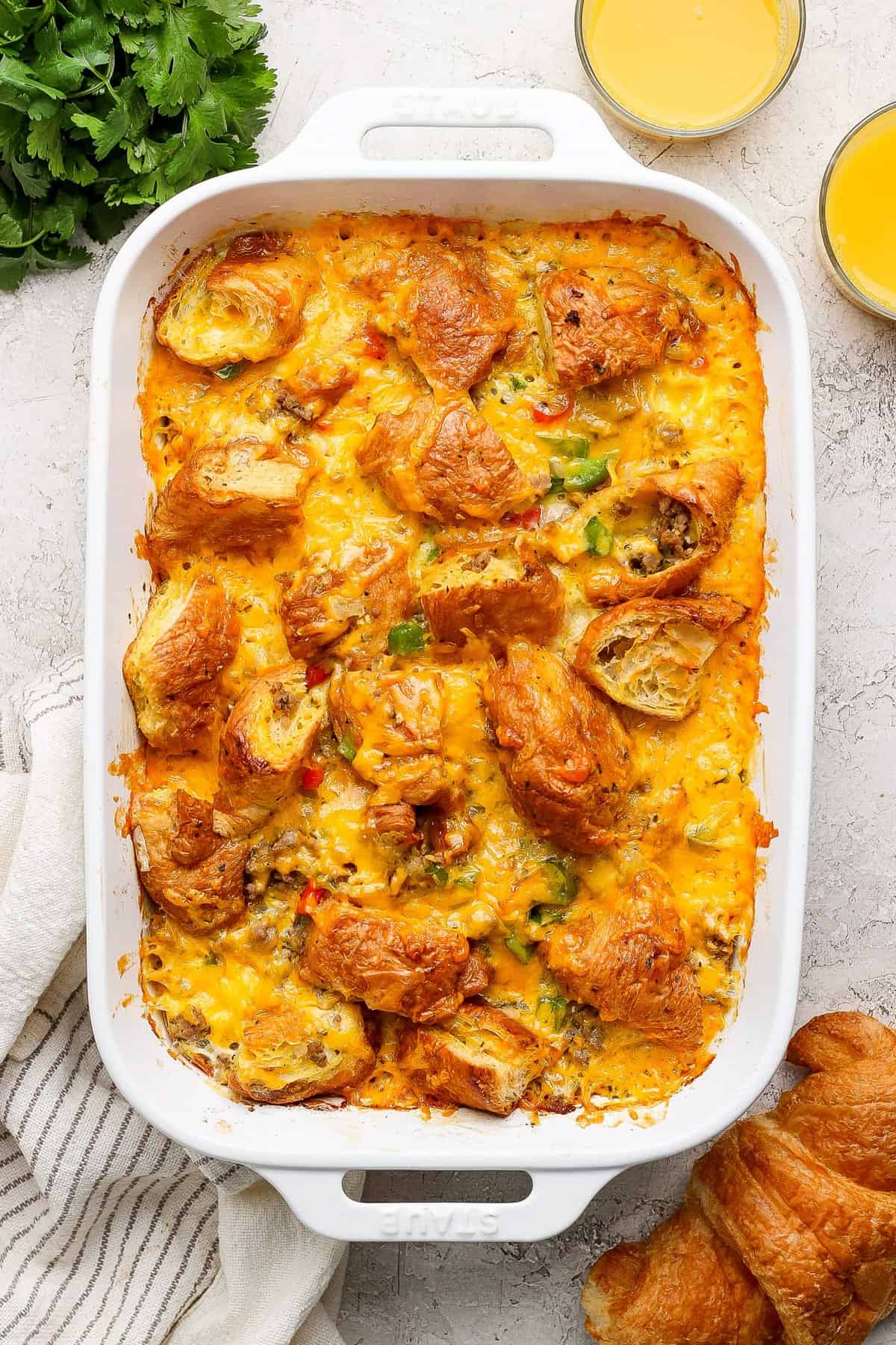 A baked casserole in a white dish, topped with melted cheese and croissant pieces. Two glasses of orange juice and fresh herbs are nearby.