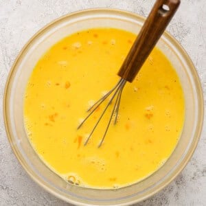 A whisk in a glass bowl containing beaten eggs on a textured surface.