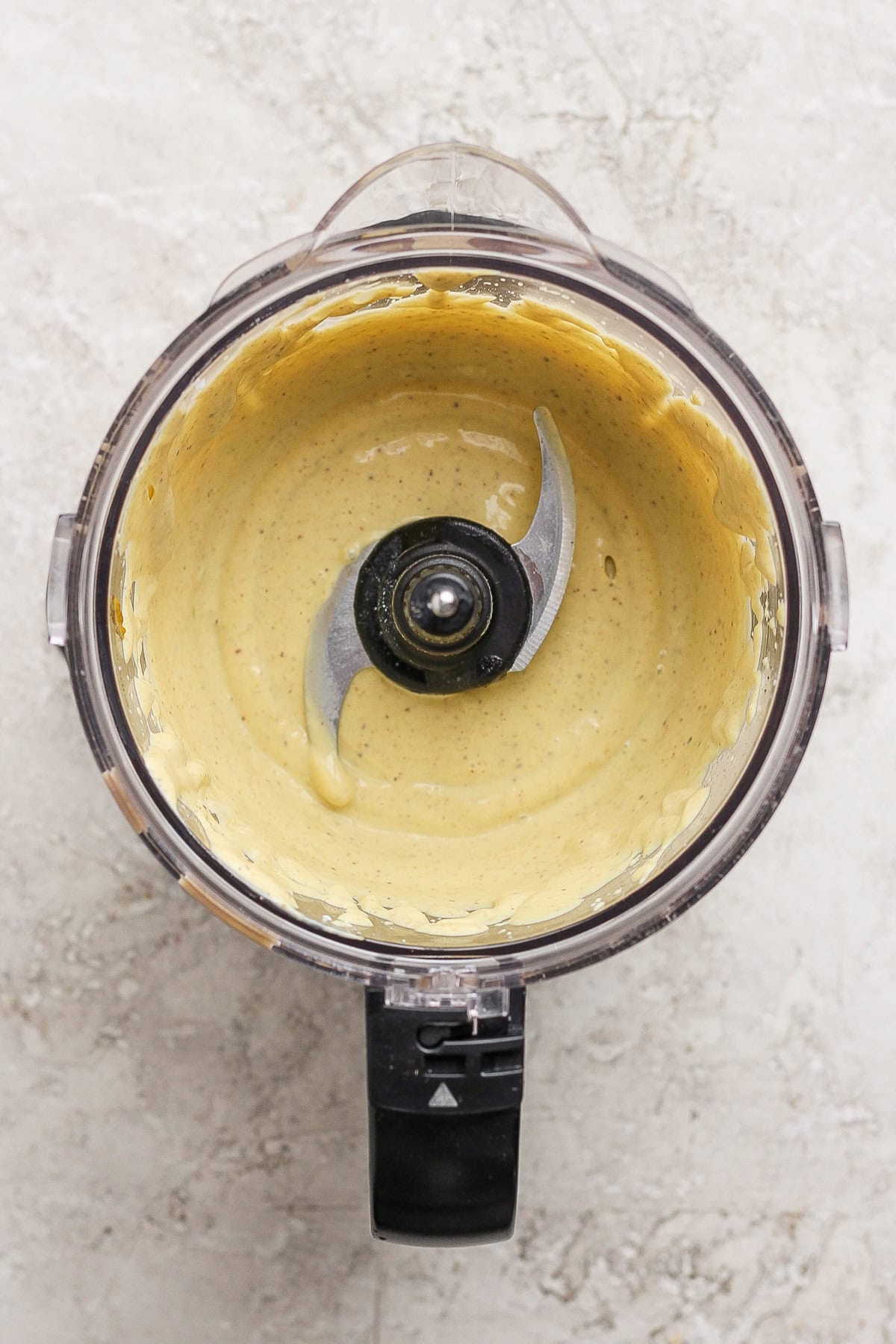 A food processor bowl containing a smooth, pale yellow creamy mixture with the blade visible in the center, set on a light textured surface.