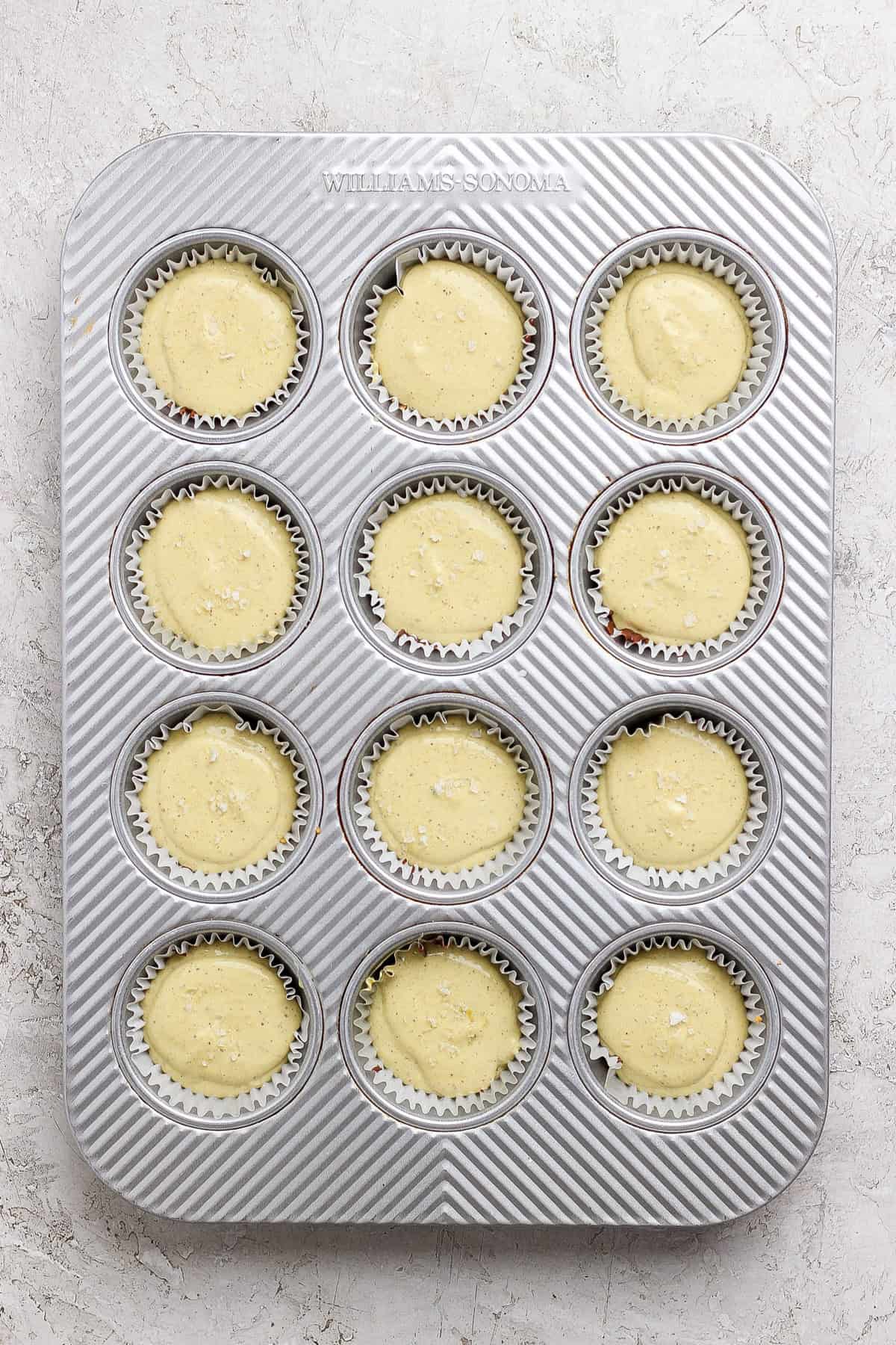 A metal muffin tin filled with twelve paper liners, each containing unbaked cupcake batter, sits on a light textured surface.