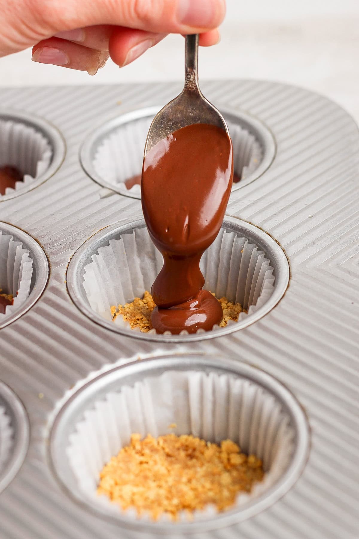 A hand holds a spoon pouring melted chocolate over crushed graham crackers in a paper-lined muffin tin.