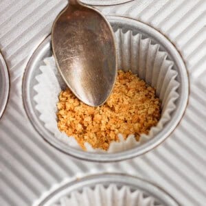 A spoon presses crushed graham cracker crumbs into a paper-lined muffin tin.