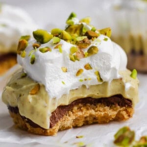 A dessert bar with a crumbly base, chocolate layer, creamy filling, whipped topping, and chopped pistachios, with a bite taken out of it.