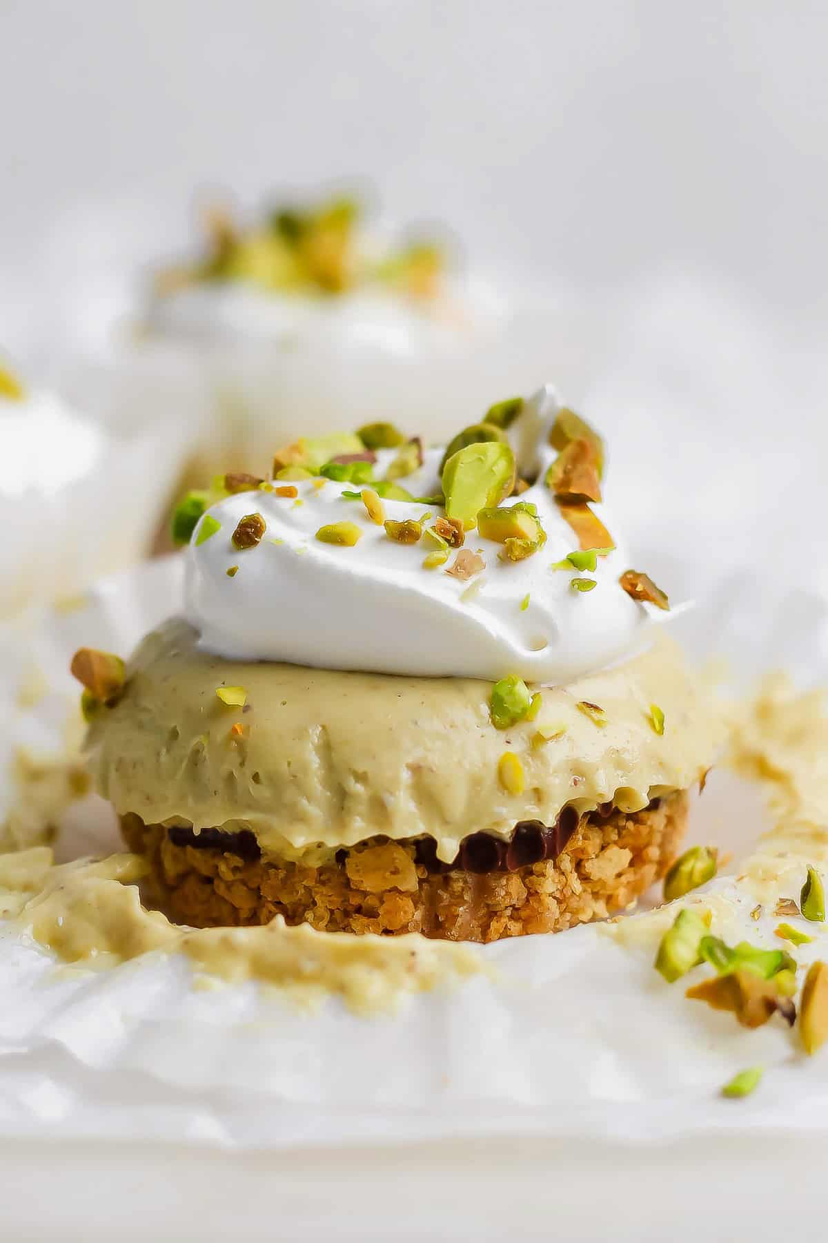 A mini cheesecake with a crumb crust, topped with whipped cream and chopped pistachios, sits on a piece of white parchment paper.
