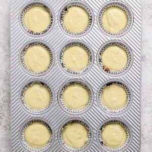 A metal muffin tin filled with twelve paper liners, each containing unbaked cupcake batter, sits on a light textured surface.