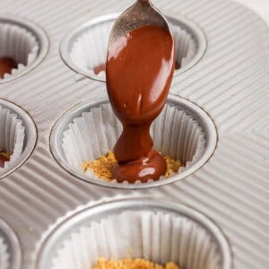 A hand holds a spoon pouring melted chocolate over crushed graham crackers in a paper-lined muffin tin.