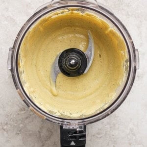 A food processor bowl containing a smooth, pale yellow creamy mixture with the blade visible in the center, set on a light textured surface.