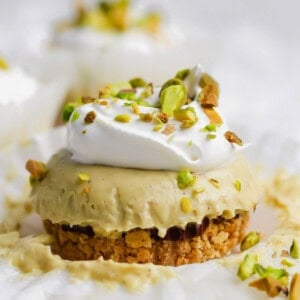 A mini cheesecake with a crumb crust, topped with whipped cream and chopped pistachios, sits on a piece of white parchment paper.