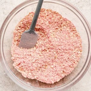 A glass bowl filled with pink-tinted oats and a gray spatula resting inside, placed on a light textured surface.