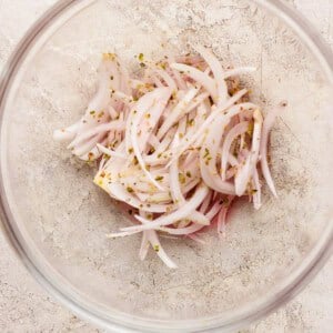 A clear glass bowl containing thinly sliced red onions mixed with dried herbs on a light, textured surface.