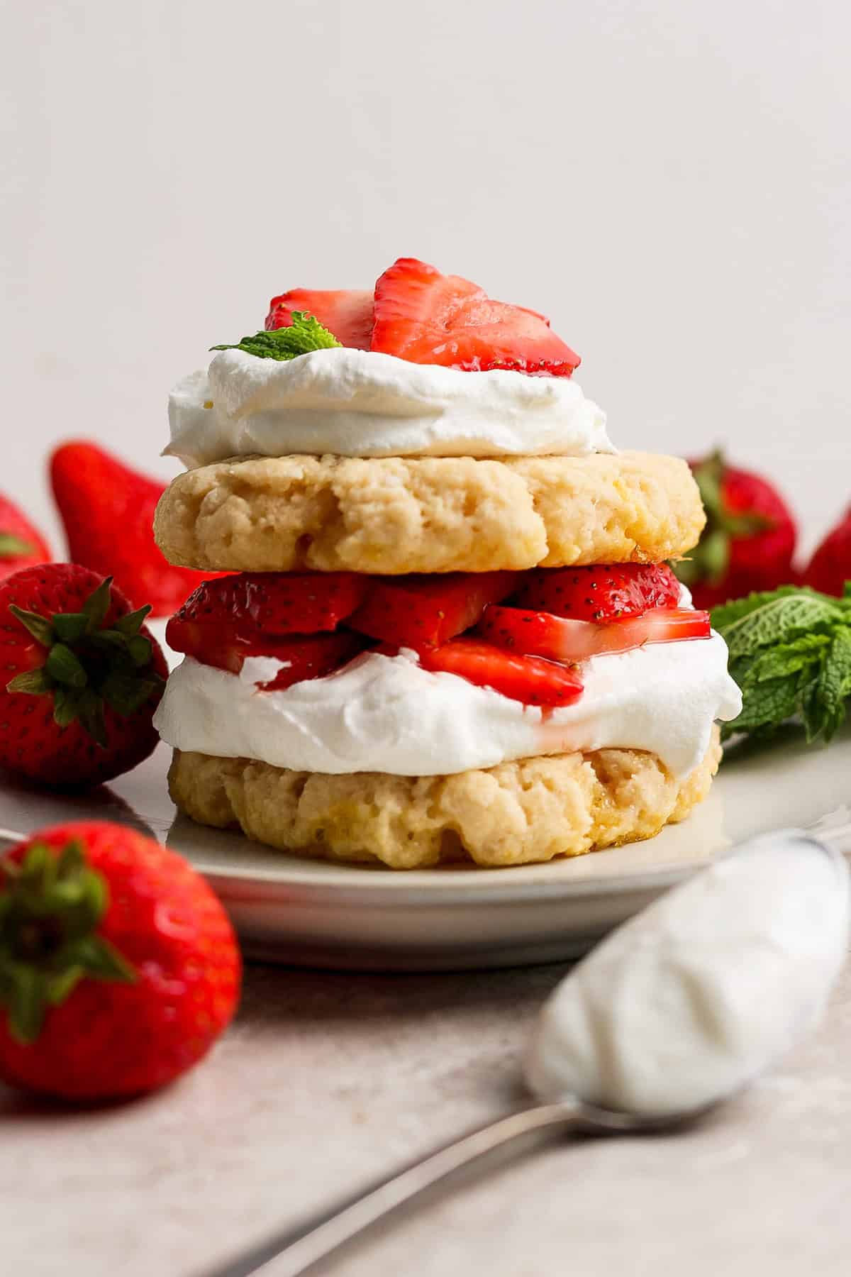 A strawberry shortcake dessert with layers of biscuits, whipped cream, and sliced strawberries on a plate, surrounded by whole strawberries and mint leaves.