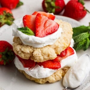 A strawberry shortcake dessert with layers of biscuits, whipped cream, and sliced strawberries, garnished with mint leaves on a white plate.