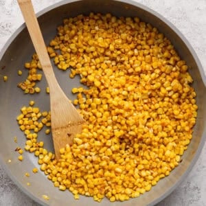 A skillet filled with sautéed corn kernels, perfect for a fresh Mediterranean corn salad, and a wooden spatula on a light gray surface.