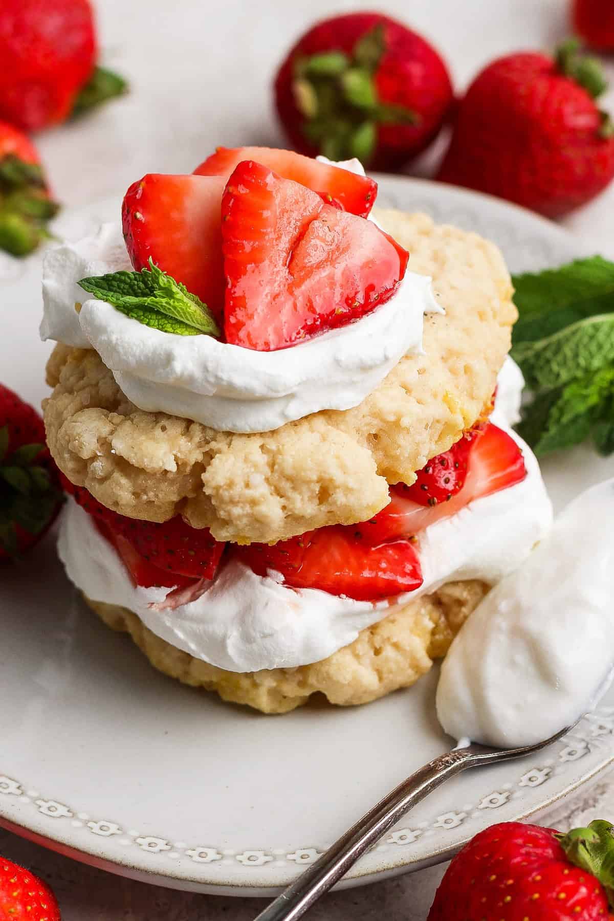 A strawberry shortcake dessert with layers of biscuits, whipped cream, and fresh strawberries, garnished with a mint leaf, on a white plate with a spoon and whole strawberries.