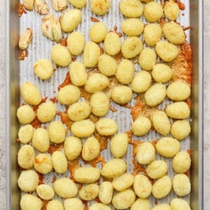 A metal baking sheet with scattered cooked gnocchi, some clumps of melted cheese, and a few roasted garlic cloves on a textured surface.