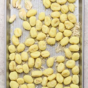 A metal baking tray with uncooked gnocchi, shredded cheese, and garlic cloves arranged on a light, textured surface.