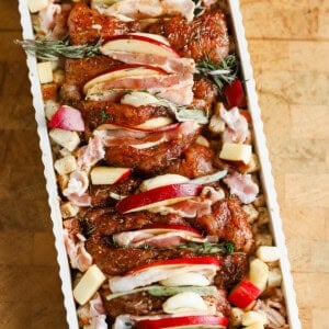 Raw seasoned chicken pieces topped with apple slices and herbs, arranged in a rectangular baking dish with cubed bread and chopped apples.