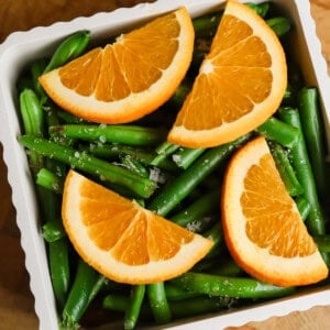 A white dish filled with green beans, topped with four orange slices, set on a wooden surface.