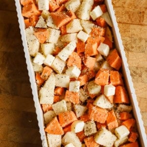 A rectangular baking dish filled with cubed sweet potatoes and white potatoes, topped with small cubes of butter and sprinkled with seasoning, on a wooden surface.