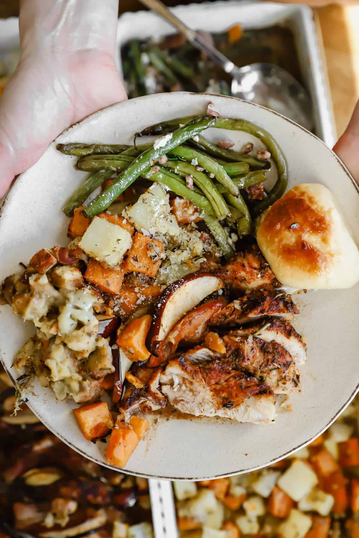 A plate with roasted turkey, green beans, stuffing, roasted vegetables, and a dinner roll, held over a table with more food trays.