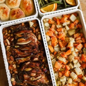 Four trays of food including dinner rolls, green beans with orange slices, roasted chicken with vegetables, and a mixed root vegetable medley on a wooden surface.