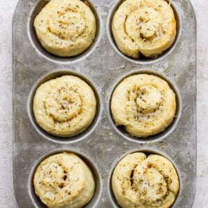 A metal muffin tray holding six unbaked, spiral-shaped dough rolls topped with herbs and shredded cheese on a light textured surface.