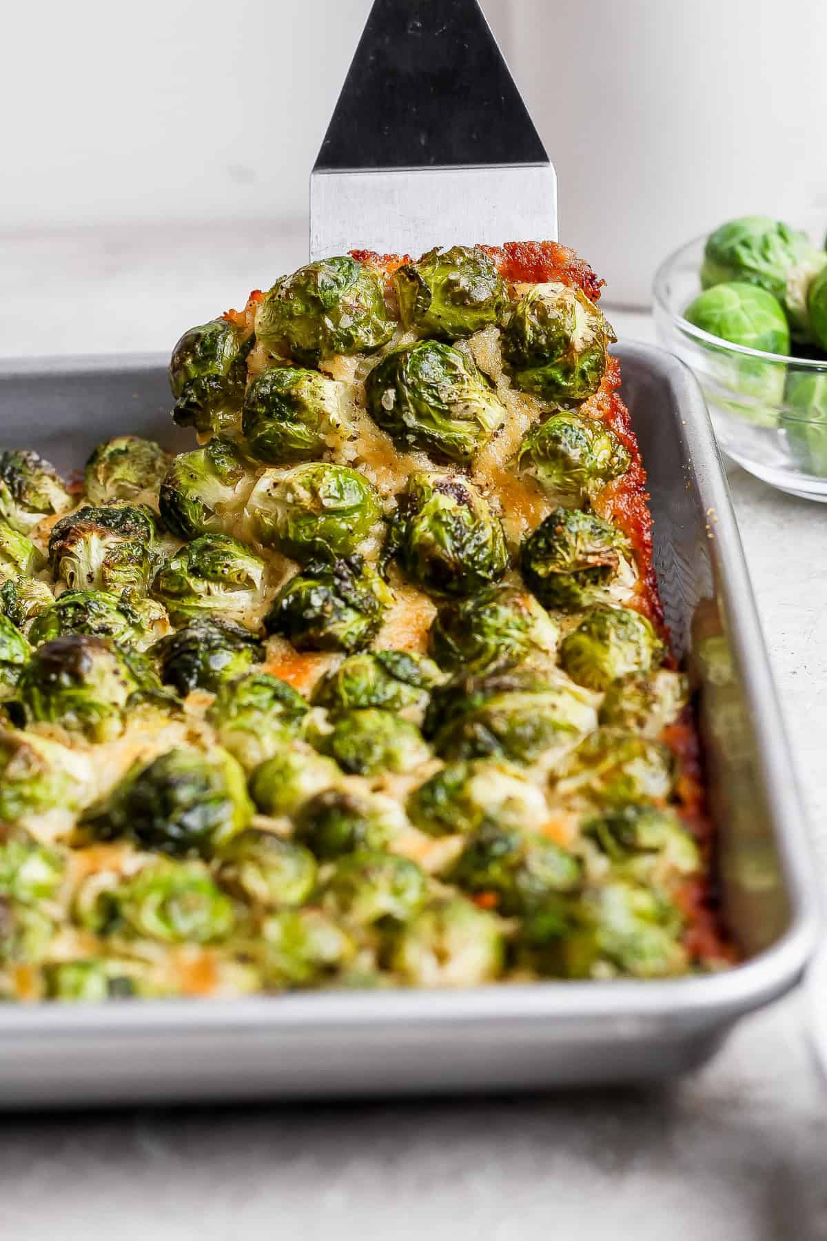 A metal spatula lifts a piece of baked casserole topped with roasted brussels sprouts from a rectangular baking pan. A bowl of raw brussels sprouts sits in the background.