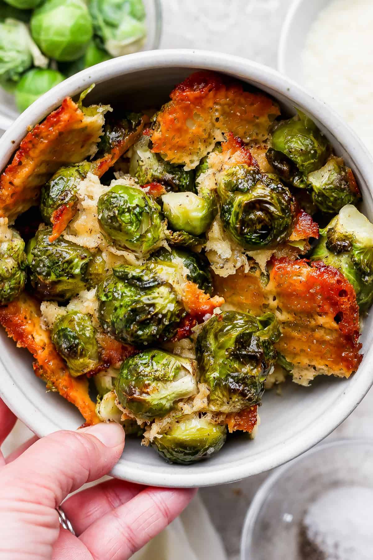 A bowl of roasted Brussels sprouts topped with crispy, golden-brown cheese, with a hand holding the bowl.