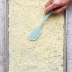 A hand uses a light blue spatula to smooth a layer of yellowish powder or crumbs on a rectangular baking sheet.
