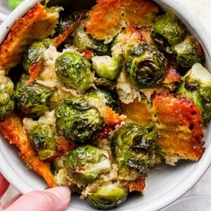 A bowl of roasted Brussels sprouts topped with crispy, golden-brown cheese, with a hand holding the bowl.