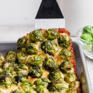 A spatula lifts a square piece of baked casserole topped with roasted Brussels sprouts from a metal pan. A bowl with raw Brussels sprouts sits in the background.