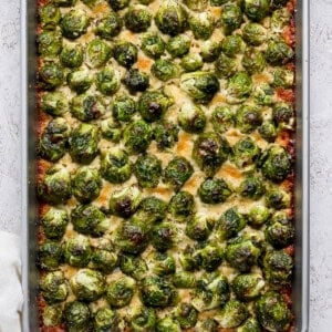 A baking sheet filled with roasted Brussels sprouts, lightly browned and topped with grated cheese. Bowls of additional Brussels sprouts and cheese are visible nearby.